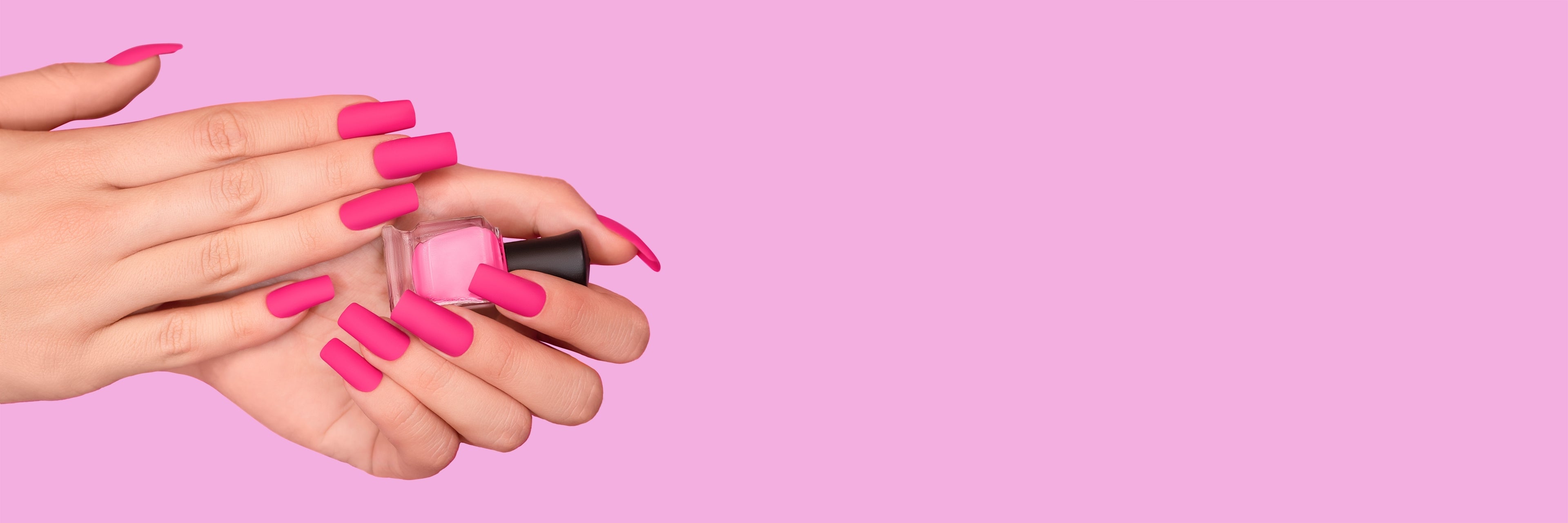 A lady's left and right hand with pink nails holding a nail polish bottle, on a light pink background.