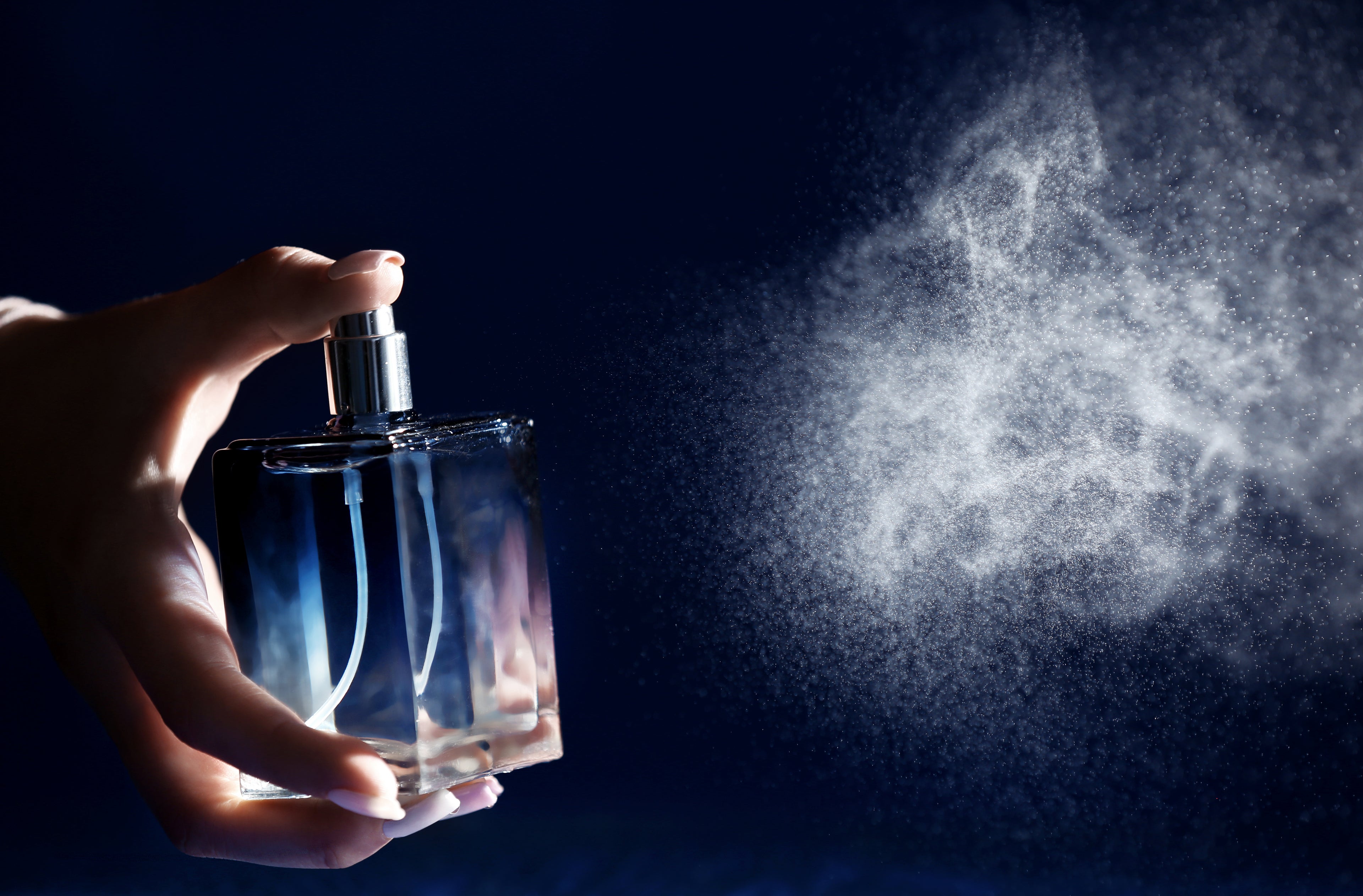 A lady spraying a bottle of perfume, with the vapour shown in a white cloud, on a dark blue background.